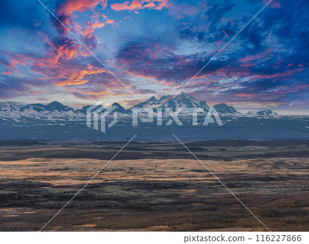 A stunning view of snow-capped peaks in the distance, contrasted by a flat, brown landscape and a vibrant blue sky with pink and purple clouds in Iceland. A stunning view of snow-capped peaks in the distance, contrasted by a flat, brown landscape and a vibrant blue sky with pink and purple clouds in Iceland. 116227866
