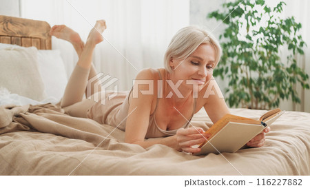 Enjoying reading. Home rest. Happy peaceful relaxed woman in sleeping suit laying with novel book on comfortable bed in light hygge interior. Enjoying reading. Home rest. Happy peaceful relaxed woman in sleeping suit laying with novel book on comfortable bed in light hygge interior. 116227882