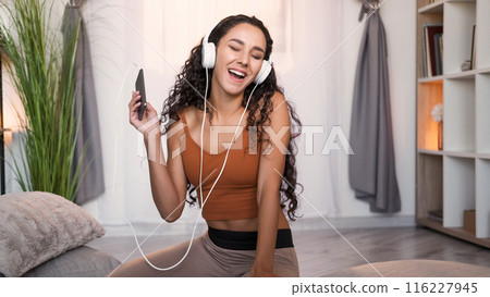Mp3 leisure. Music weekend. Happy smiling girl in headphones enjoying listening to audio on phone singing at light modern home interior with free space. Mp3 leisure. Music weekend. Happy smiling girl in headphones enjoying listening to audio on phone singing at light modern home interior with free space. 116227945