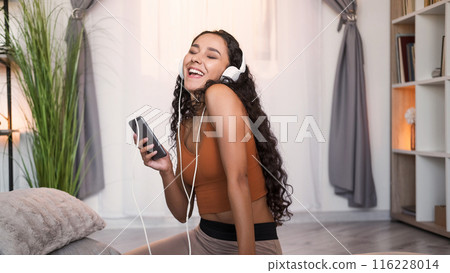 Music fun. Weekend playlist. Inspired happy girl enjoying listening to mp3 audio in headphones using mobile phone app singing at modern home interior with free space. Music fun. Weekend playlist. Inspired happy girl enjoying listening to mp3 audio in headphones using mobile phone app singing at modern home interior with free space. 116228014