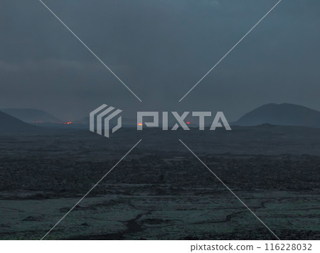 Aerial view of a dark volcanic landscape in Iceland at dusk, featuring small glowing red craters. The sky is a deep blue, contrasting with the black ground. Aerial view of a dark volcanic landscape in Iceland at dusk, featuring small glowing red craters. The sky is a deep blue, contrasting with the black ground. 116228032