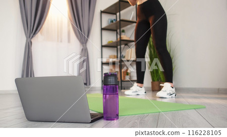 Fitness tutorial. Remote training. Online yoga group. Laptop water bottle closeup before unrecognizable woman fit legs in sportswear working out home interior. Fitness tutorial. Remote training. Online yoga group. Laptop water bottle closeup before unrecognizable woman fit legs in sportswear working out home interior. 116228105