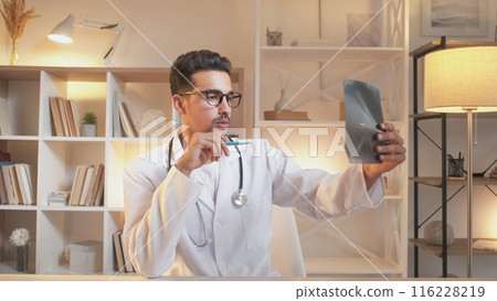 Clinical diagnosis. Traumatologist doctor. Smart surgeon man in white uniform studying x ray film sitting at modern interior clinic office workplace. Clinical diagnosis. Traumatologist doctor. Smart surgeon man in white uniform studying x ray film sitting at modern interior clinic office workplace. 116228219