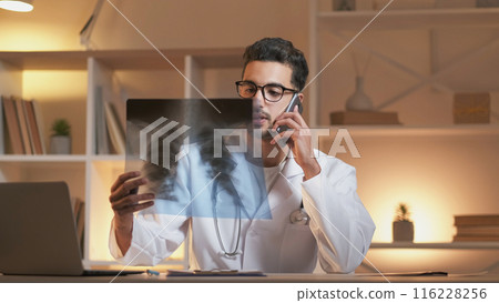 Phone diagnosis. Mobile doctor consultation. Pneumonia disease. Pulmonologist man with x ray of lungs talking on smartphone sitting at work desk in clinic interior. Phone diagnosis. Mobile doctor consultation. Pneumonia disease. Pulmonologist man with x ray of lungs talking on smartphone sitting at work desk in clinic interior. 116228256