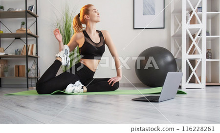 Stretch exercise. Online session. Gymnastic class. Muscle soreness relief. Free movement. Female fitness instructor in sportswear pull leg on yoga mat with laptop at home interior. Stretch exercise. Online session. Gymnastic class. Muscle soreness relief. Free movement. Female fitness instructor in sportswear pull leg on yoga mat with laptop at home interior. 116228261