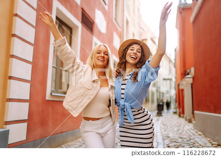 Two women are walking down a street, one wearing a white jacket and the other wearing a blue shirt and white skirt. They are smiling and holding hands, giving off a happy and friendly vibe 116228672