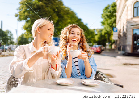 Portrait of a smiling young women friends sitting outdoors in cafe drinking coffee. Beauty, relax. 116228735