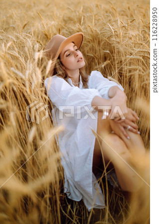 Young woman in white linen dress and hat enjoying a sunny day in a golden wheat field. Summer, beauty. 116228950