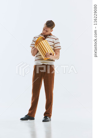 Young man dressed in retro-style clothes, polo shirt and pants, looking inside cardboard present box with interest, isolated on white background. Young man dressed in retro-style clothes, polo shirt and pants, looking inside cardboard present box with interest, isolated on white background. 116229080