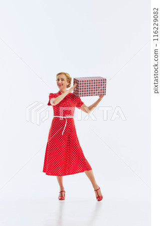 Elegant woman in red polka dot dress holds large checked gift box with cheerful smile. Moment of celebration and joy. Elegant woman in red polka dot dress holds large checked gift box with cheerful smile. Moment of celebration and joy. 116229082