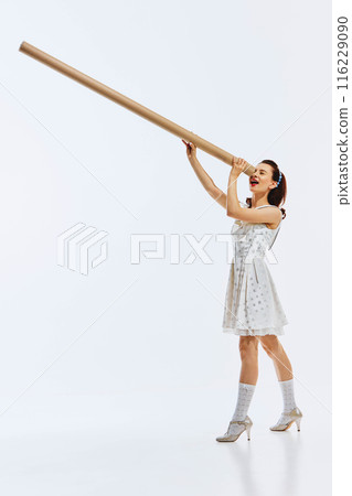 Young woman in polka dot dress and vintage shoes looking into giant cardboard tube isolated on white background. Looking for innovation and inspiration. 116229090
