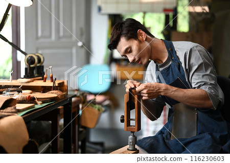 Skilled craftsman in apron sewing while making handmade leather wallet in workshop Skilled craftsman in apron sewing while making handmade leather wallet in workshop 116230603