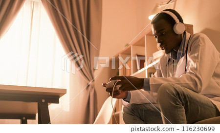Doctor break. Digital leisure. Mobile entertainment. Male physician in headphones enjoying playing video game online on phone at modern home interior with copy space. Doctor break. Digital leisure. Mobile entertainment. Male physician in headphones enjoying playing video game online on phone at modern home interior with copy space. 116230699