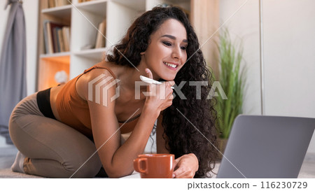 Internet research. Online learning. Distance education. Curious cheerful girl watching video lesson using laptop taking notes at modern home interior with copy space. 116230729