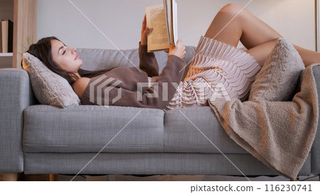 Home reading. Literature enjoying. Indoor weekend. Pleased peaceful young relaxing woman resting with book on comfortable couch in cozy living room. Home reading. Literature enjoying. Indoor weekend. Pleased peaceful young relaxing woman resting with book on comfortable couch in cozy living room. 116230741
