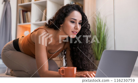 Online chat. Virtual leisure. Internet communication. Curious woman typing using laptop on floor at home living room interior with free space. Online chat. Virtual leisure. Internet communication. Curious woman typing using laptop on floor at home living room interior with free space. 116230759