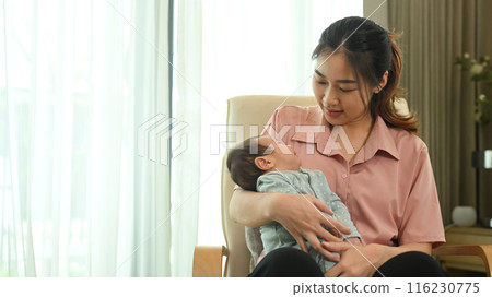 Young mother looking tenderly at her sleeping infant in a cozy light blue onesie. Motherhood and child care concept 116230775