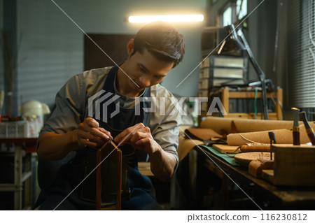 Professional craftsman sewing while making handmade leather wallet in workshop 116230812