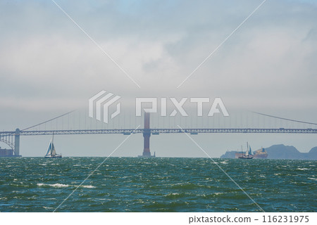 Iconic Golden Gate Bridge in mist, San Francisco. View from water shows bridge amidst choppy bay waters with sailboats. Overcast sky, hills in background. Shows natural beauty and engineering marvel. Iconic Golden Gate Bridge in mist, San Francisco. View from water shows bridge amidst choppy bay waters with sailboats. Overcast sky, hills in background. Shows natural beauty and engineering marvel. 116231975