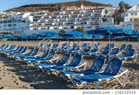 Beach sunbeds with blue umbrellas, chairs on sandy beach on Tenerife seaside resort, coastal town leisure 116232014