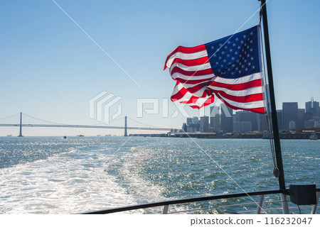 American flag fluttering on boat as it leaves SF towards Bay Bridge. Skyline, buildings, river wake visible on sunny day. Maritime culture essence captured. 116232047