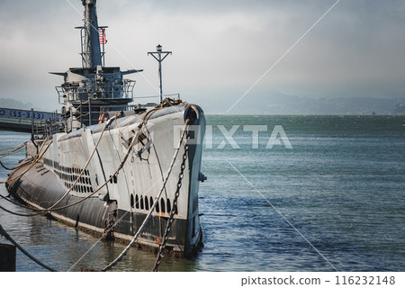 Large submarine moored at pier in calm bay waters. Hull number visible, tied with ropes. Overcast sky gives serene ambiance. Possibly San Francisco Bay area. perfect for tourists. 116232148