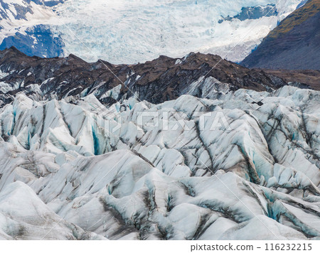 Stunning aerial view of a glacier in Iceland, showcasing snow and ice with exposed rock, surrounded by majestic mountains under a clear blue sky. 116232215