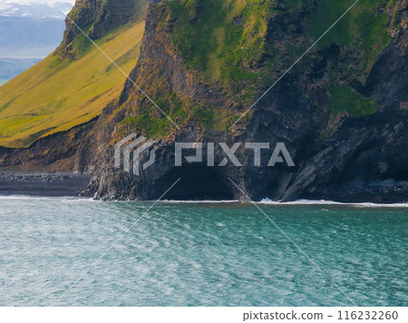 A stunning aerial view of Iceland's black sand beach featuring a large cave in a cliff face made of basalt columns. Deep blue water and light blue sky complete the scene. A stunning aerial view of Iceland's black sand beach featuring a large cave in a cliff face made of basalt columns. Deep blue water and light blue sky complete the scene. 116232260