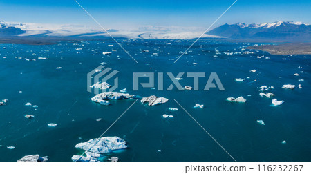 A breathtaking aerial view of calm blue waters with ice floes, reflecting a clear sky, and distant snow-capped mountains in Iceland. 116232267