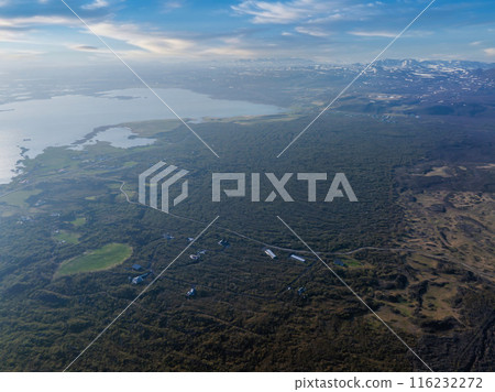 High vantage point captures a vast green landscape in Iceland, featuring a winding road and a distant body of water, likely a lake or fjord. High vantage point captures a vast green landscape in Iceland, featuring a winding road and a distant body of water, likely a lake or fjord. 116232272