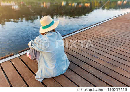 Back view woman wearing straw hat denim jacket sit on wooden pier dock river or lake gazing calm water during peaceful moment. Alone sad female person thinking at pond jetty Back view woman wearing straw hat denim jacket sit on wooden pier dock river or lake gazing calm water during peaceful moment. Alone sad female person thinking at pond jetty 116237526