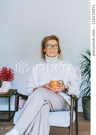 Middle aged woman relaxing with pumpkin shaped cup of hot drink in scandy style hygge interior home with fall mood decor. Lady dreaming, enjoy calm mood without stress, well being alone. Cozy autumn 116238851