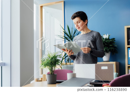 Motivated newcomer middle aged neutral gender female standing in office unpacking box with personal belongings, checking project plan at workplace having first working day concept. New Carrier start 116238890
