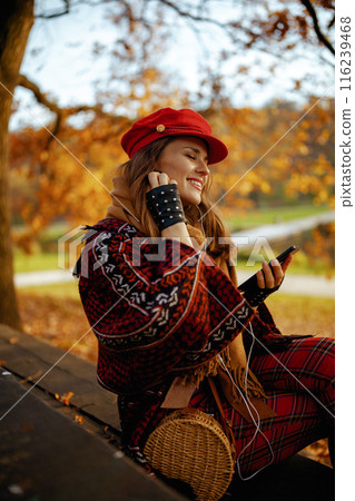 smiling woman using phone while sitting on bench in city park 116239468