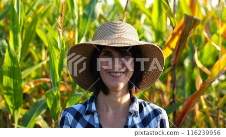 Happy smiling female farmer looks into camera standing near corn field. Portrait of adult beautiful agronomist in straw hat with maize meadow at background. Agricultural business concept. Close up 116239956
