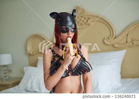 Alluring woman in leather mask sexually eating banana on bed 116240071