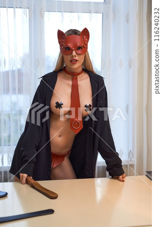 Bossy woman in red bondage and mask with paddle standing behind the table Bossy woman in red bondage and mask with paddle standing behind the table 116240232