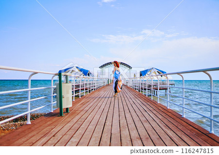 With the sea as a captivating backdrop, a red-haired woman in a summer dress and backpack graces the resort pier with her presence, creating a scene of timeless beauty. With the sea as a captivating backdrop, a red-haired woman in a summer dress and backpack graces the resort pier with her presence, creating a scene of timeless beauty. 116247815