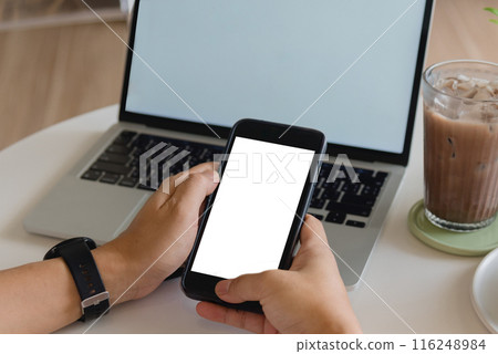 Hands holding blank-screen smartphone in front of laptop on white table, modern remote work setting. 116248984