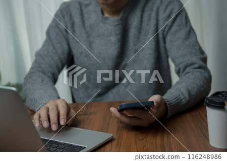 Man in gray sweater using a laptop and holding a smartphone at a wooden table with a coffee cup, focusing on multitasking in a work-from-home setup. 116248986