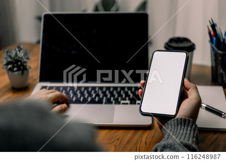 Person using a smartphone while sitting at a desk with a laptop, suggesting multitasking or mobile communication. The screen of the smartphone is blank. 116248987