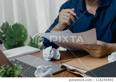 Person reviewing papers at a desk with a laptop and crumpled paper balls 116248989