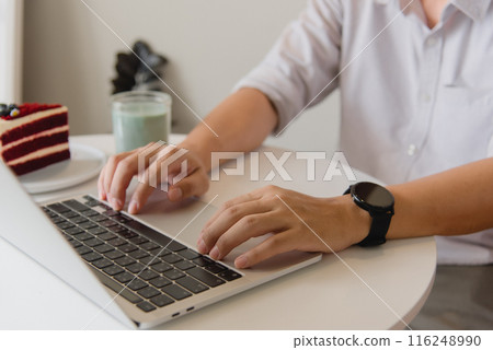 Hands typing on a laptop with a slice of cake on a white table, creating a cozy work environment. 116248990