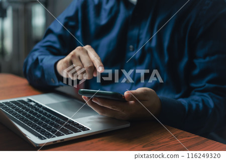 Close-up of a businessperson multitasking with a mobile phone and laptop at a wooden desk Close-up of a businessperson multitasking with a mobile phone and laptop at a wooden desk 116249320