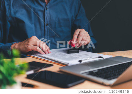 A person's hands are seen stamping a document, symbolizing the approval process in a professional setting. The laptop in the background suggests a modern office environment. 116249346