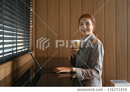 Young businesswoman holding paper cup and using digital tablet in modern office Young businesswoman holding paper cup and using digital tablet in modern office 116251858
