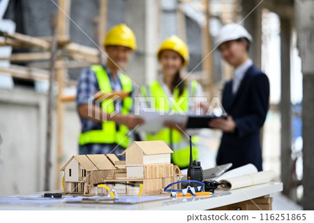 An architectural model, blueprints and tools on table with construction workers standing in blurred background 116251865
