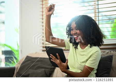 Excited young woman reading good news on digital tablet and celebrating success on couch 116251891