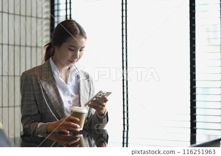 Portrait of Asian businesswoman in formalwear holding paper cup and texting on a mobile app 116251963
