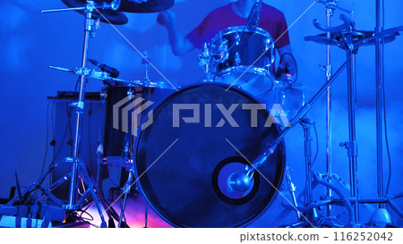 A drummer is captured in the midst of a performance on stage, surrounded by a drum kit with various cymbals and drumsticks in hand, under the bright lights of the venue. A drummer is captured in the midst of a performance on stage, surrounded by a drum kit with various cymbals and drumsticks in hand, under the bright lights of the venue. 116252042